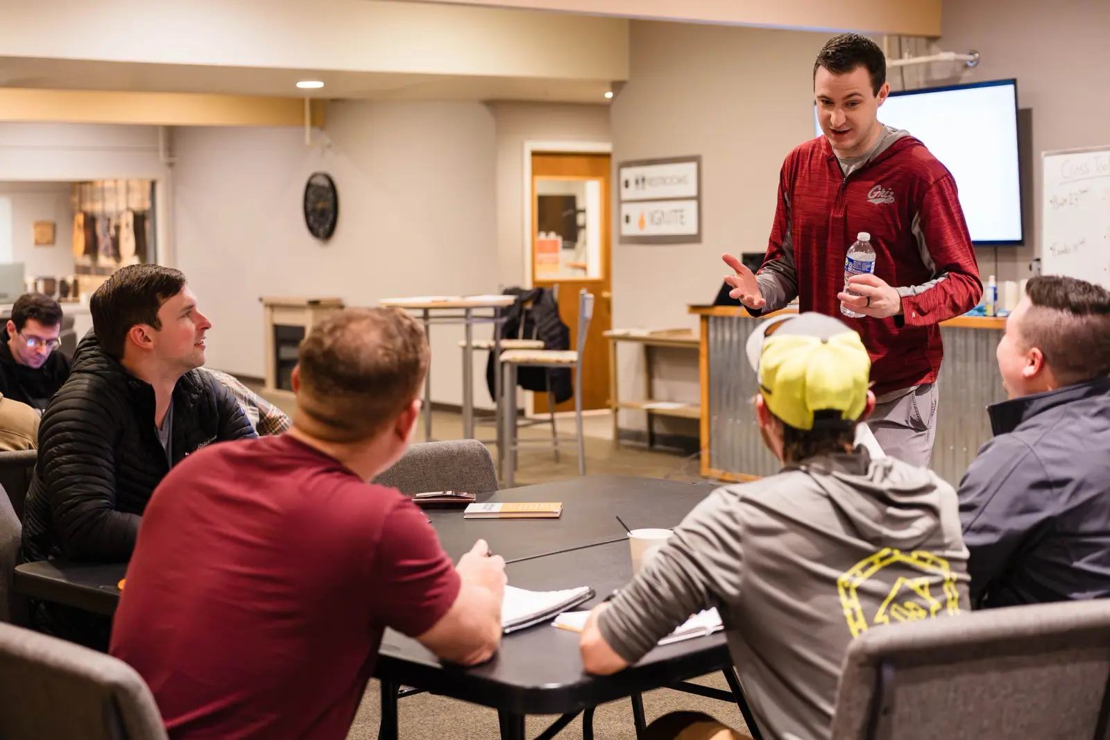 A man speaks to a small group of men seated around a table in a meeting room.