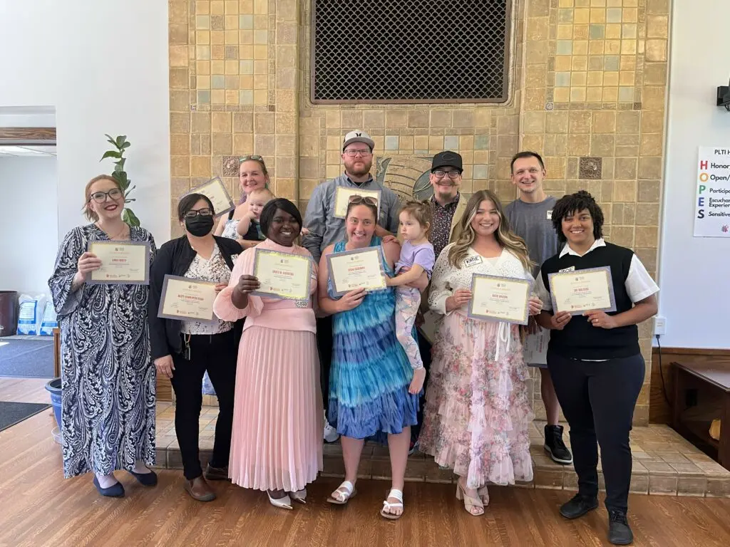 A group of adults, some with children, stand together holding certificates in a room with a decorative fireplace.