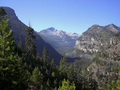 A scenic view of a mountain valley with forest-covered slopes and snow-dusted peaks in the distance.