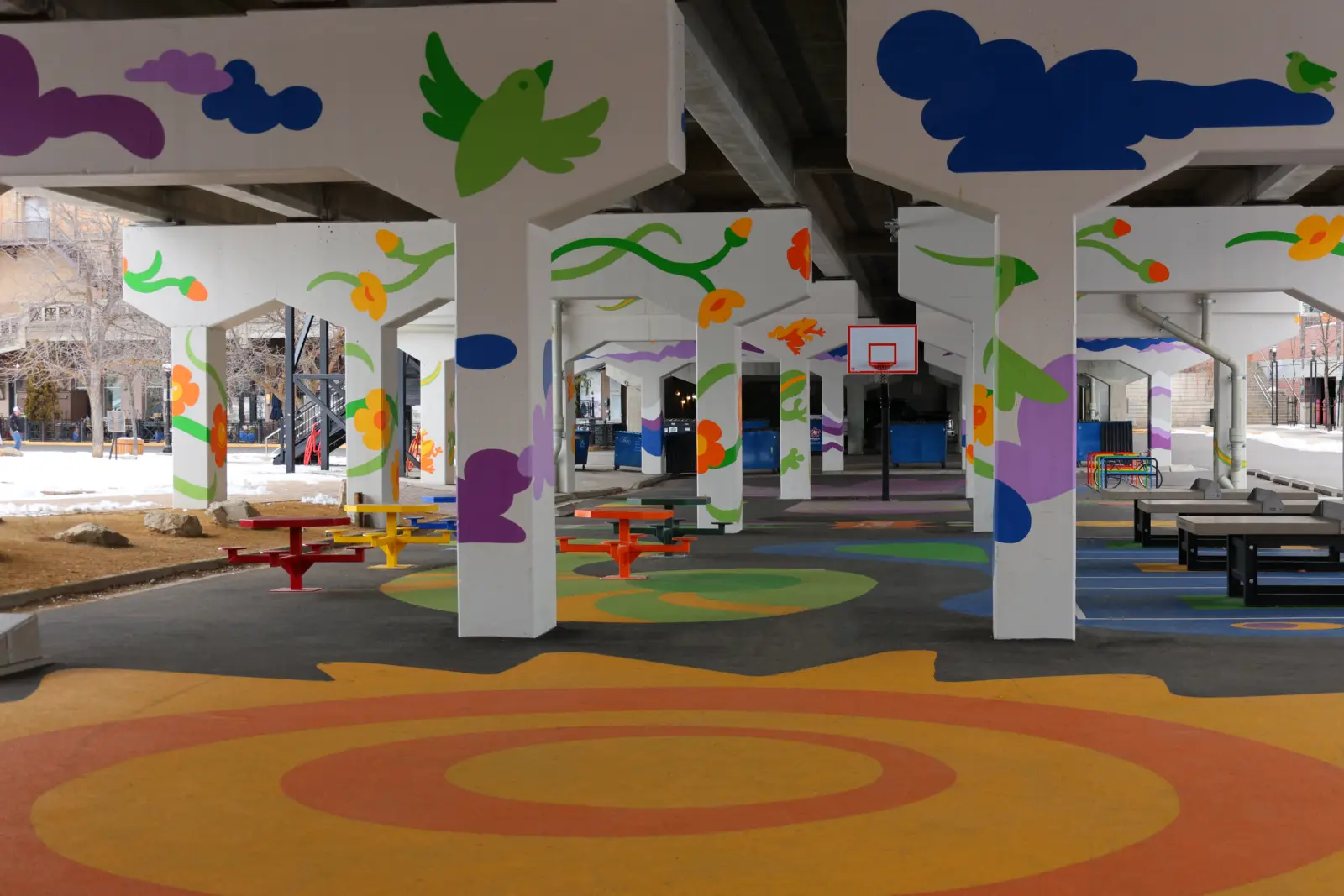 Colorful murals decorate the concrete support pillars and ground of an urban recreation area under a highway bridge.