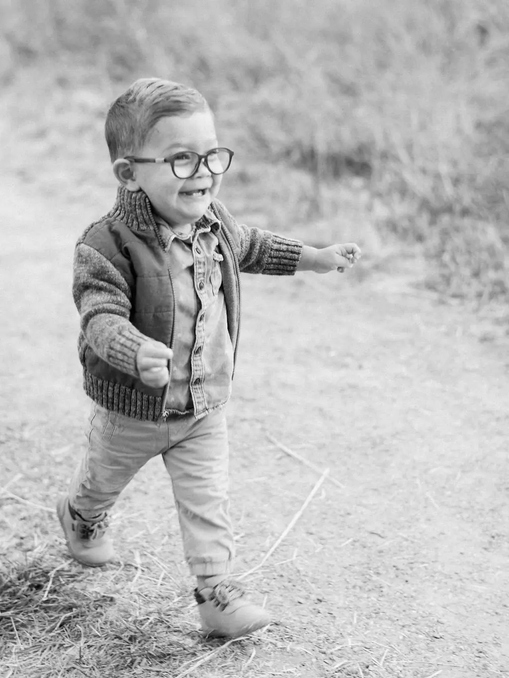 A young child wearing glasses and a jacket walks outdoors while smiling.