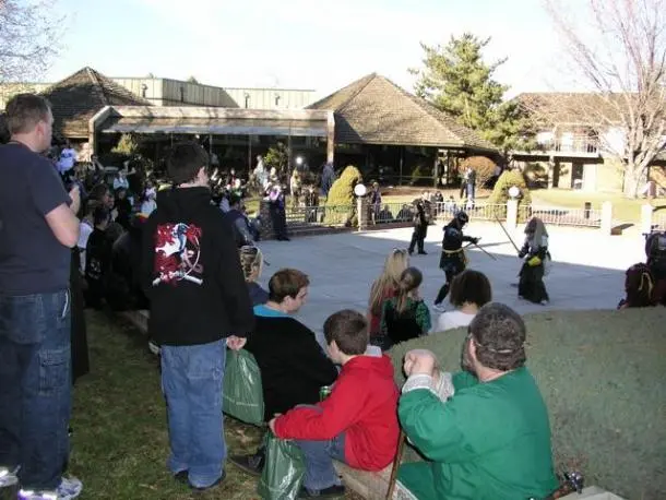 A crowd of people watches a live demonstration of stage combat outdoors in front of a building.