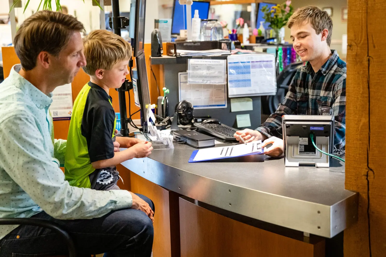 A father and son stand at a desk while a staff member helps them with paperwork.