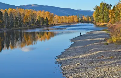 A person fishes along the rocky bank of a calm river surrounded by autumn trees.