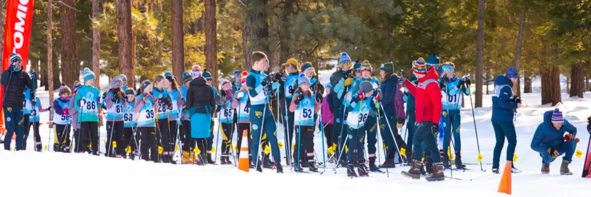 A large group of children and instructors are gathered on a snowy course, preparing for a skiing event.