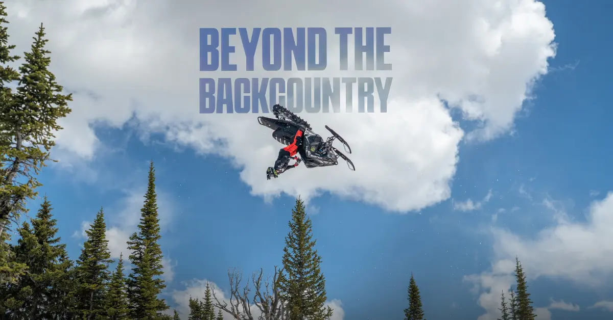 A snowmobiler performs a stunt in the air above pine trees against a blue sky.