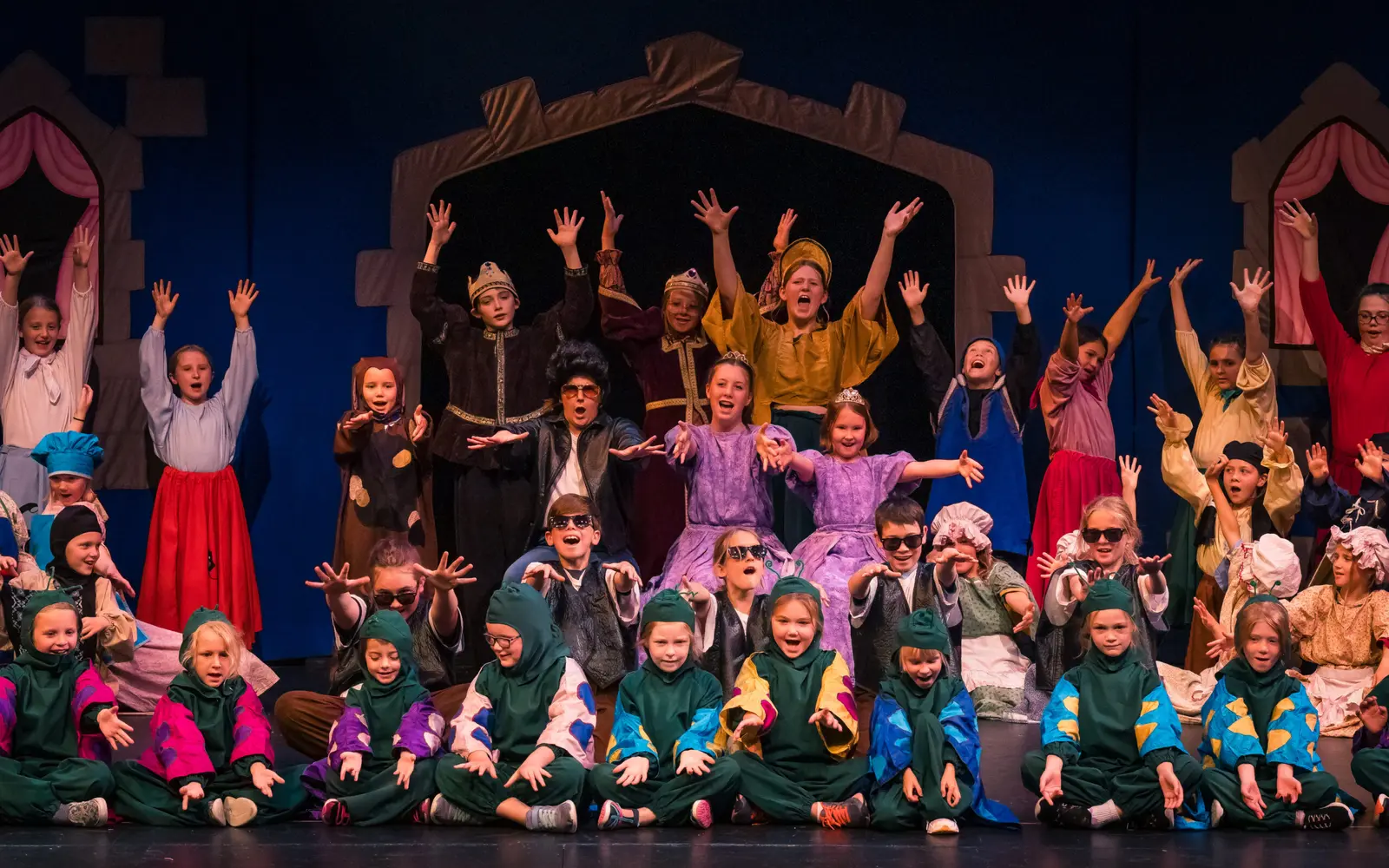 A group of children in costumes are performing on stage with their arms raised.