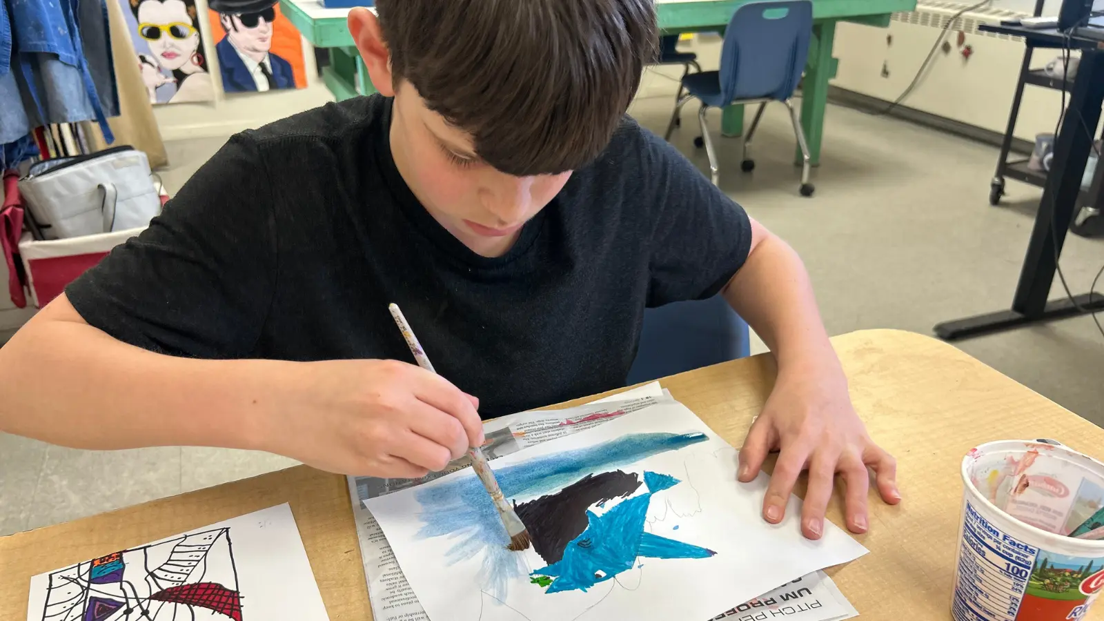 A young person sits at a desk and paints a picture of a shark on a piece of paper.