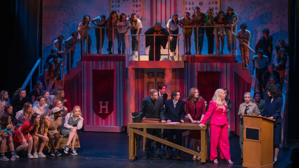 A large cast of students performs a theatrical scene on a set featuring a multi-level structure.
