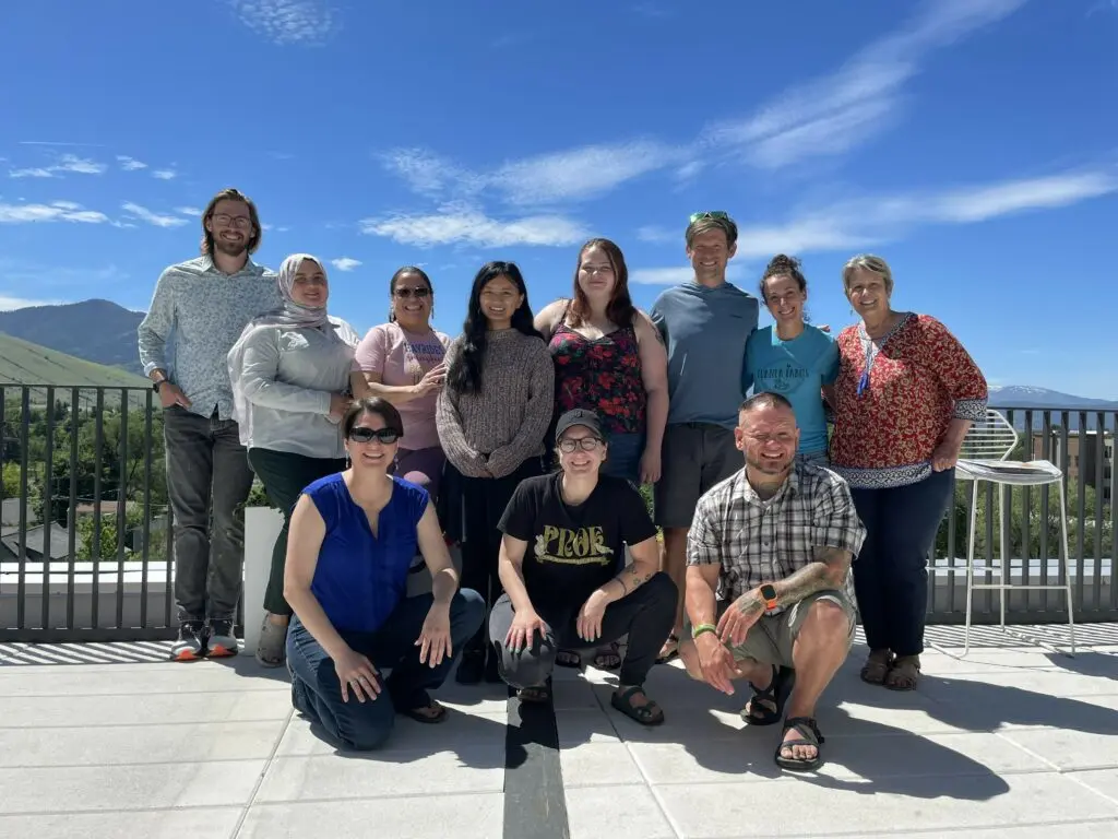 A diverse group of adults poses for a photograph on a rooftop deck under a clear blue sky.