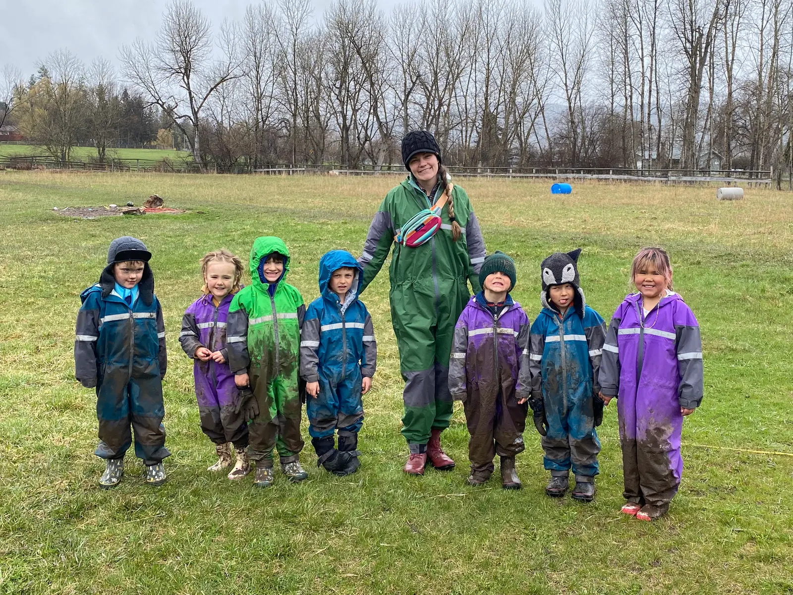 An instructor poses with a group of young children wearing muddy outdoor gear in a grassy field.