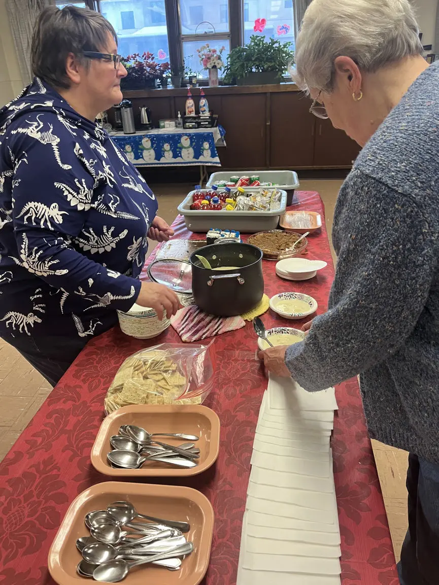 Two women serve soup and crackers from a table covered in a red tablecloth.