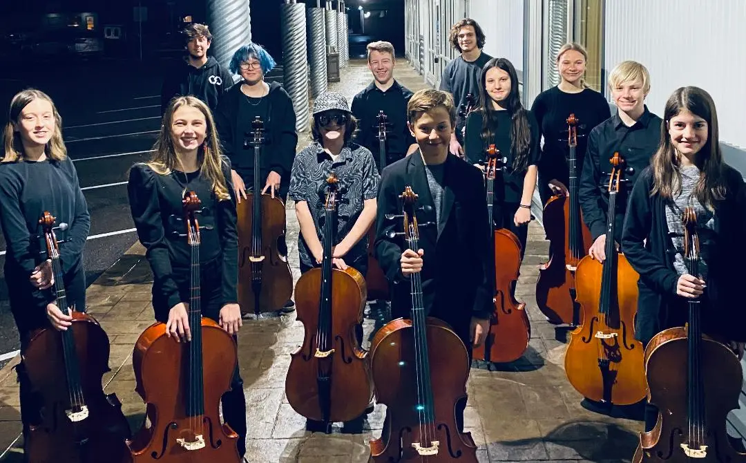 A group of young musicians poses with their cellos outside at night.