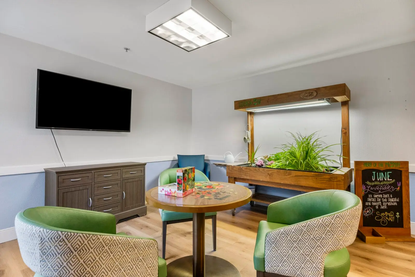 A bright room features a seating area with green chairs, a table with a puzzle, and a plant display.