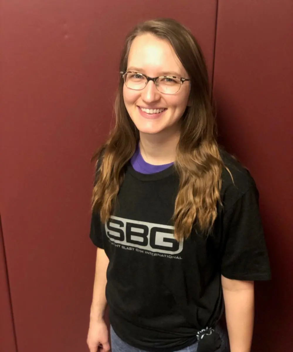 A smiling young woman wearing an SBG t-shirt stands in front of a maroon wall.