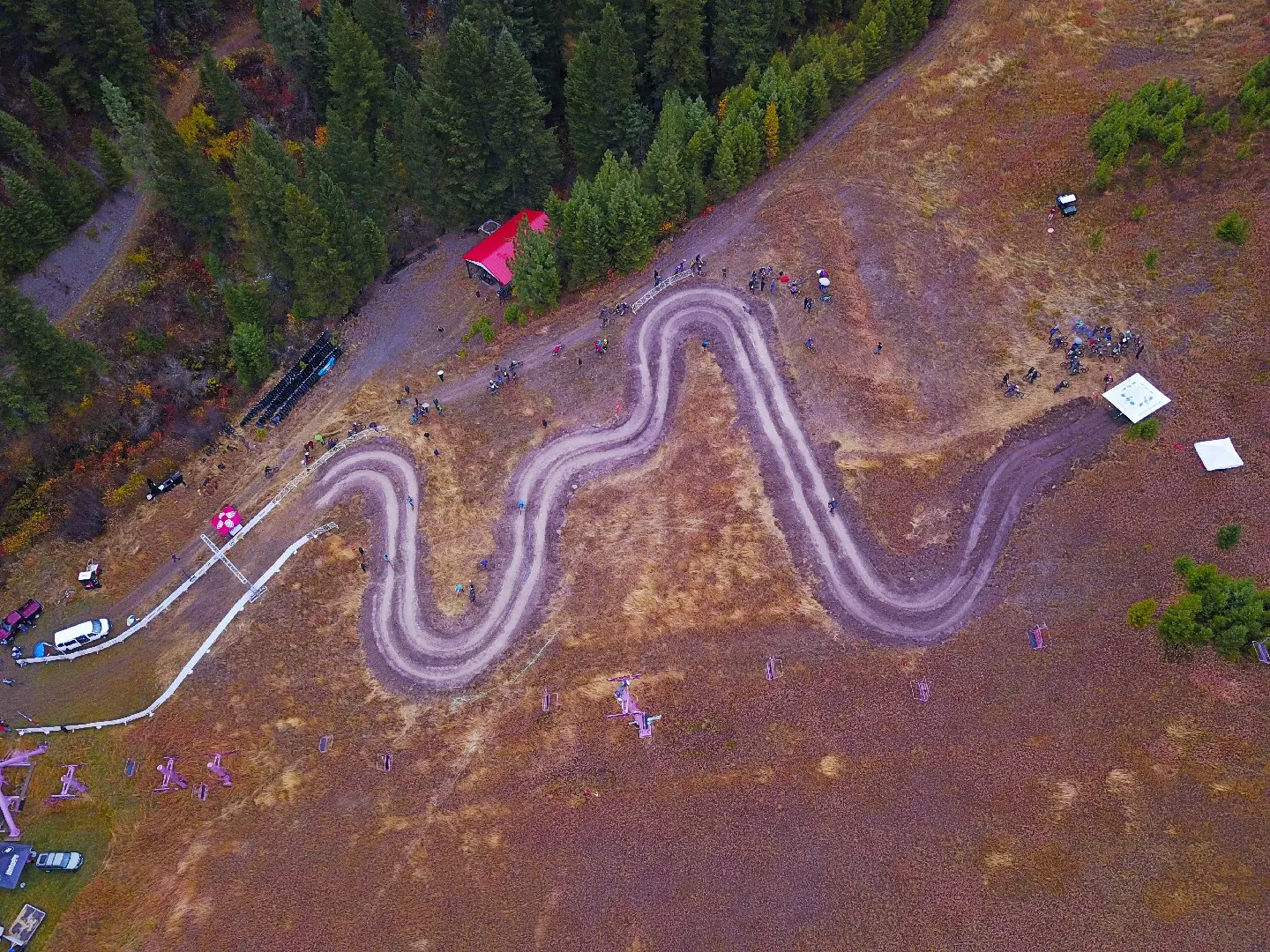 An aerial view shows cyclists riding on a serpentine dirt track on a hillside near trees and some event structures.