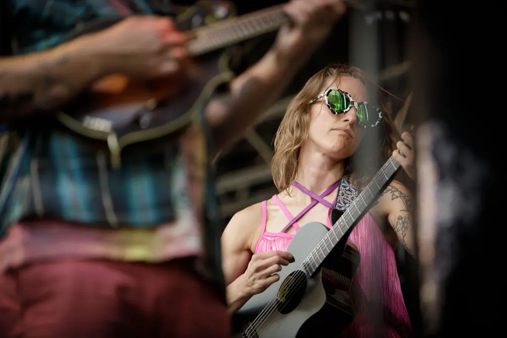 A musician wearing green sunglasses plays a guitar while performing on stage.
