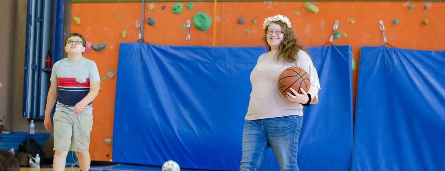 A child and an adult with a flower crown hold a basketball in front of a climbing wall.