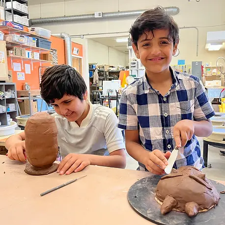 Two children work on clay sculptures at a table in an art studio.