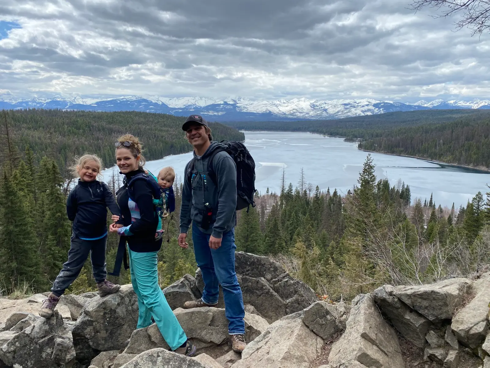 A family stands on a rocky outcrop overlooking a large lake and snow-capped mountains.