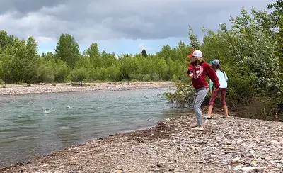 Photo from fvlt.org Two children stand on a rocky riverbank and throw stones into the water.