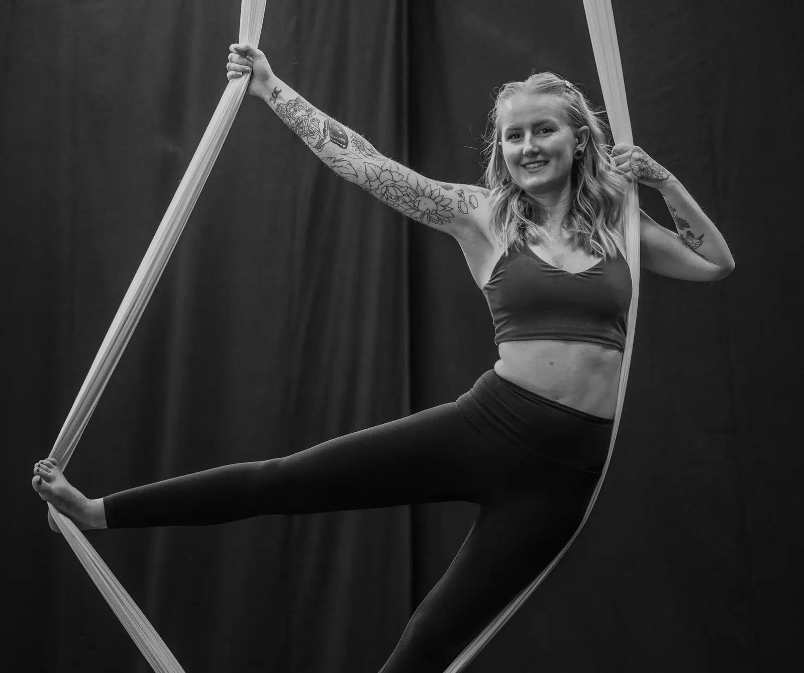 A woman performs a pose on aerial silks against a dark background.