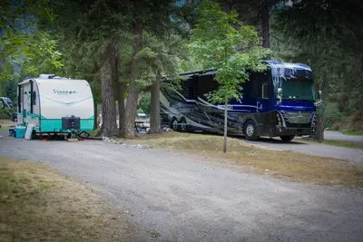Photo from campgroundstregisrvpark.com A vintage travel trailer and a large Class A motorhome parked side-by-side on spacious shaded pull-through sites among tall pine trees at Campground St. Regis