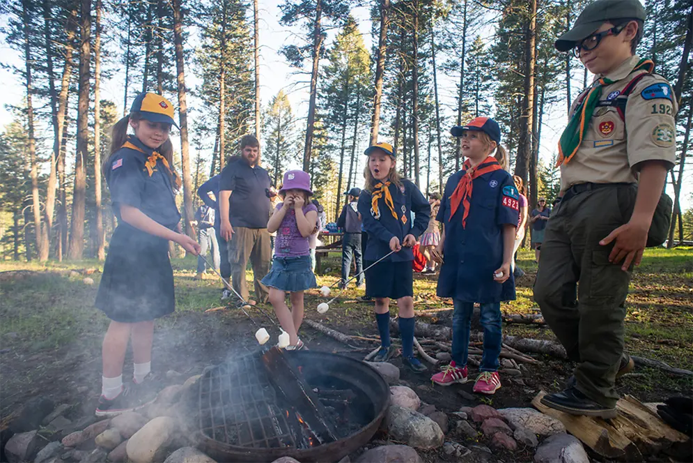 A group of children and an adult are roasting marshmallows over a campfire in the woods.