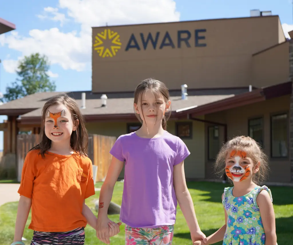 Three children with face paint stand outdoors holding hands in front of a building.