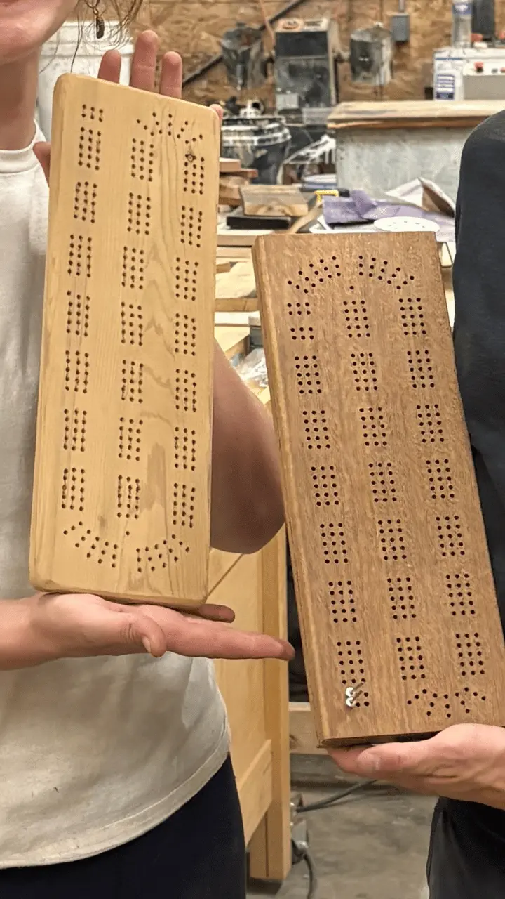 Two people hold handcrafted wooden cribbage boards in a workshop.