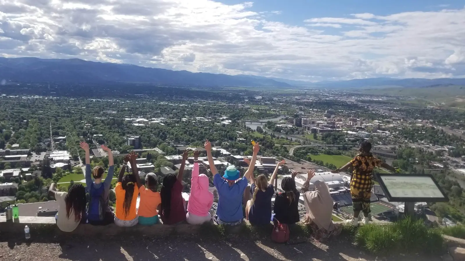 A group of young people sit with their arms raised, overlooking a city from a high vantage point.