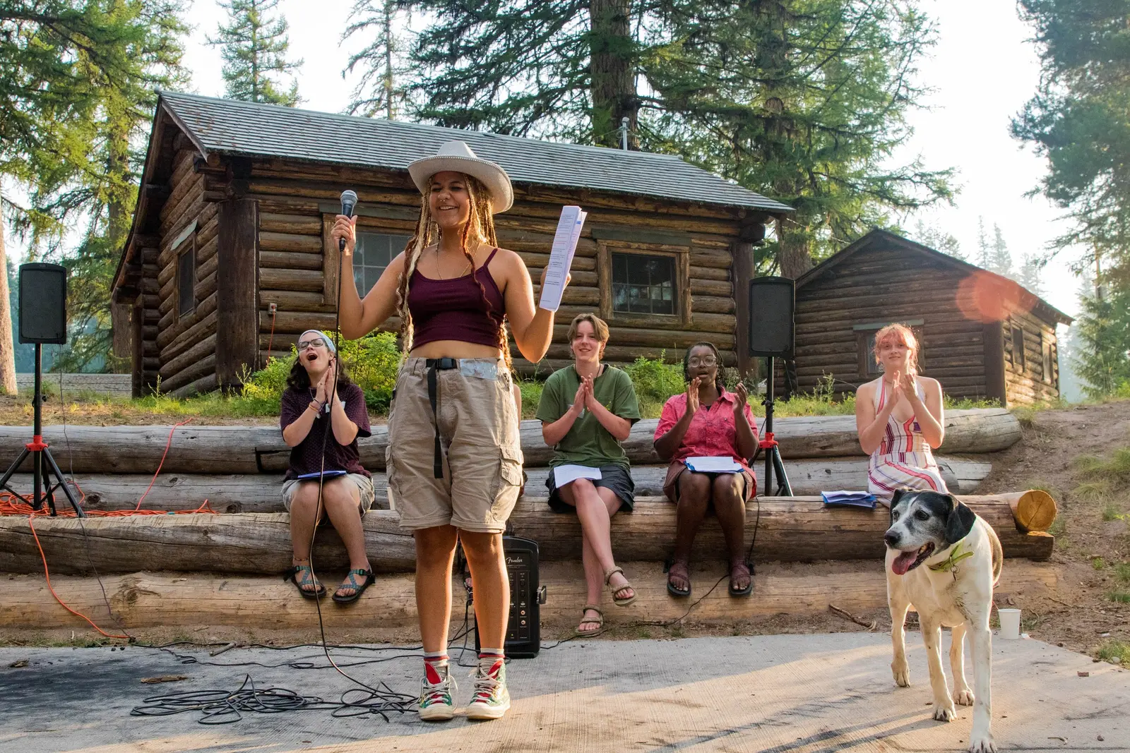 A young woman with a microphone stands in front of seated audience members outdoors near log cabins.