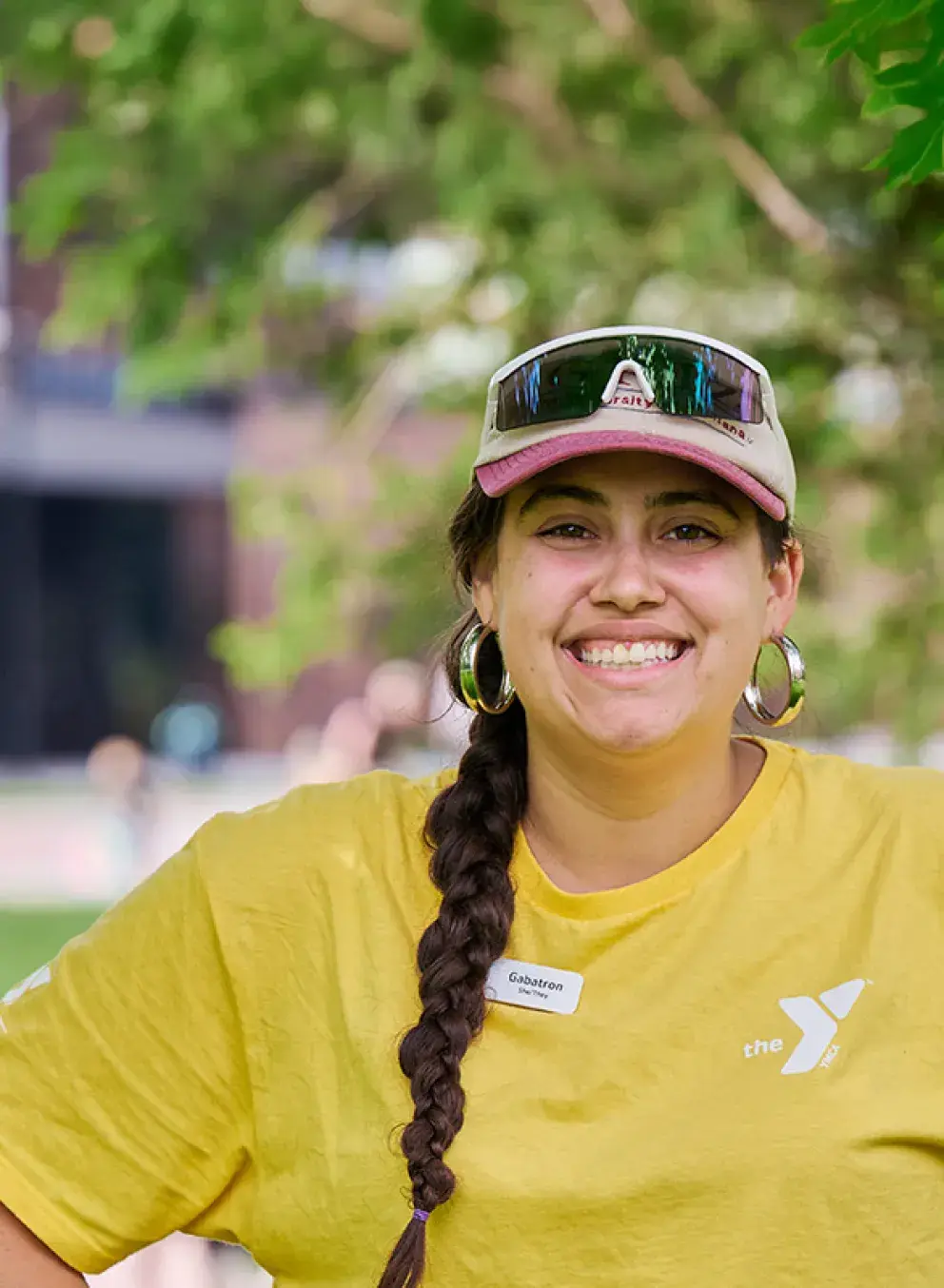 missoula ymca camp counselor wearing a yellow shirt, baseball hat, and sunglasses. they have long, brown hair in a braid.
