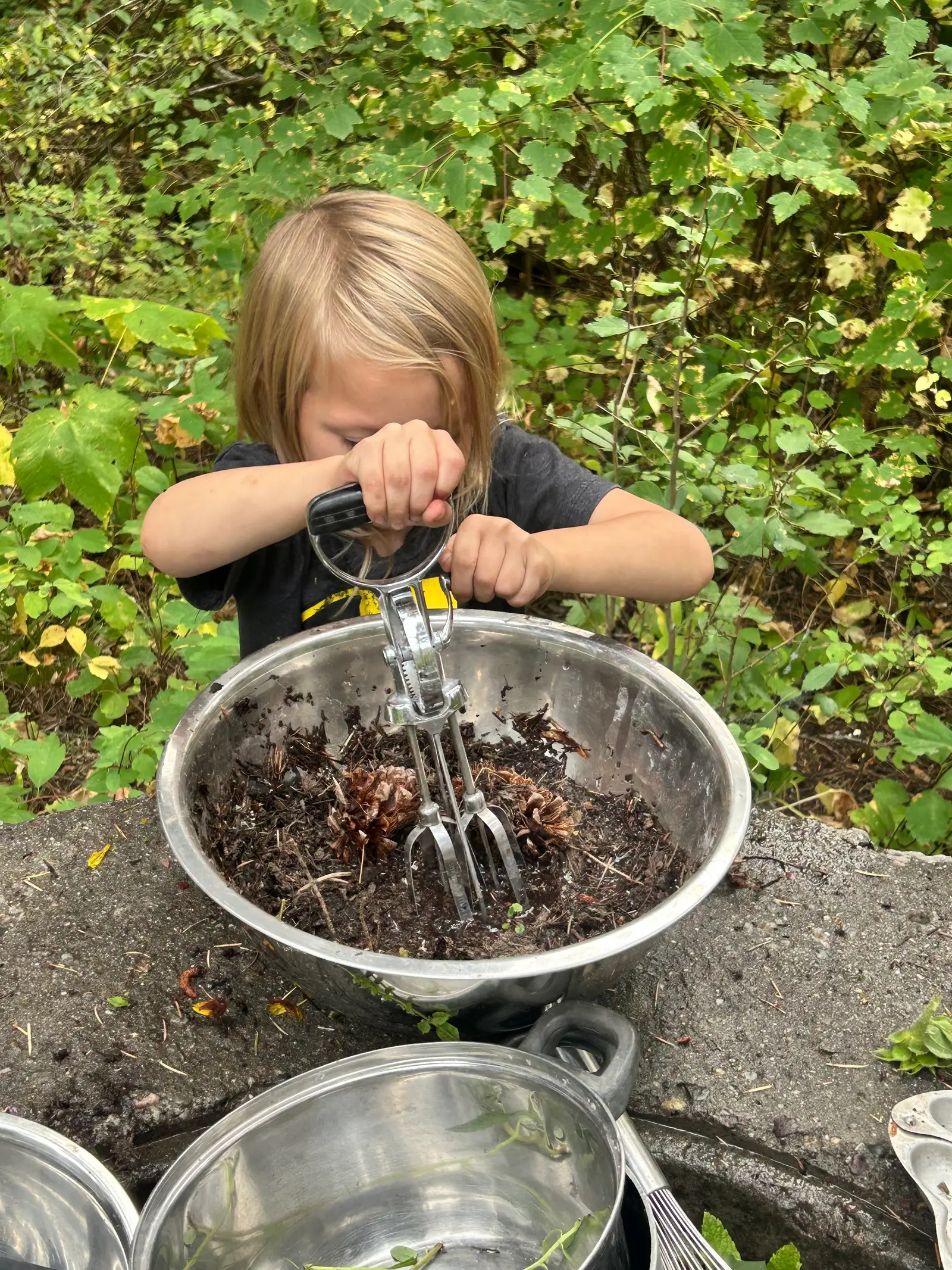 A child plays outdoors with dirt and pinecones in a mixing bowl.