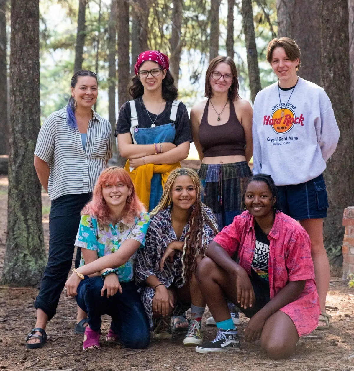 A diverse group of seven young people pose for a photo together in a wooded area.