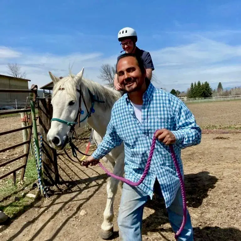 A person leads a horse with a rider wearing a helmet in an outdoor paddock.