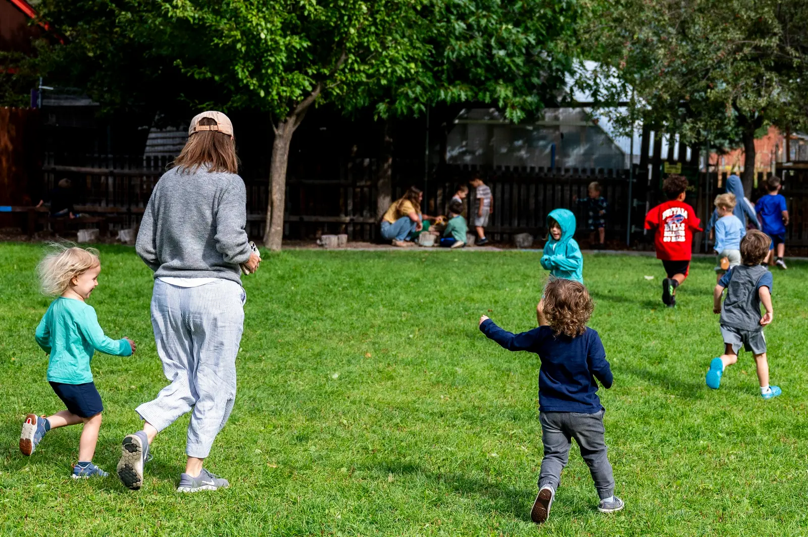 A group of children and an adult are running through a grassy yard outdoors.
