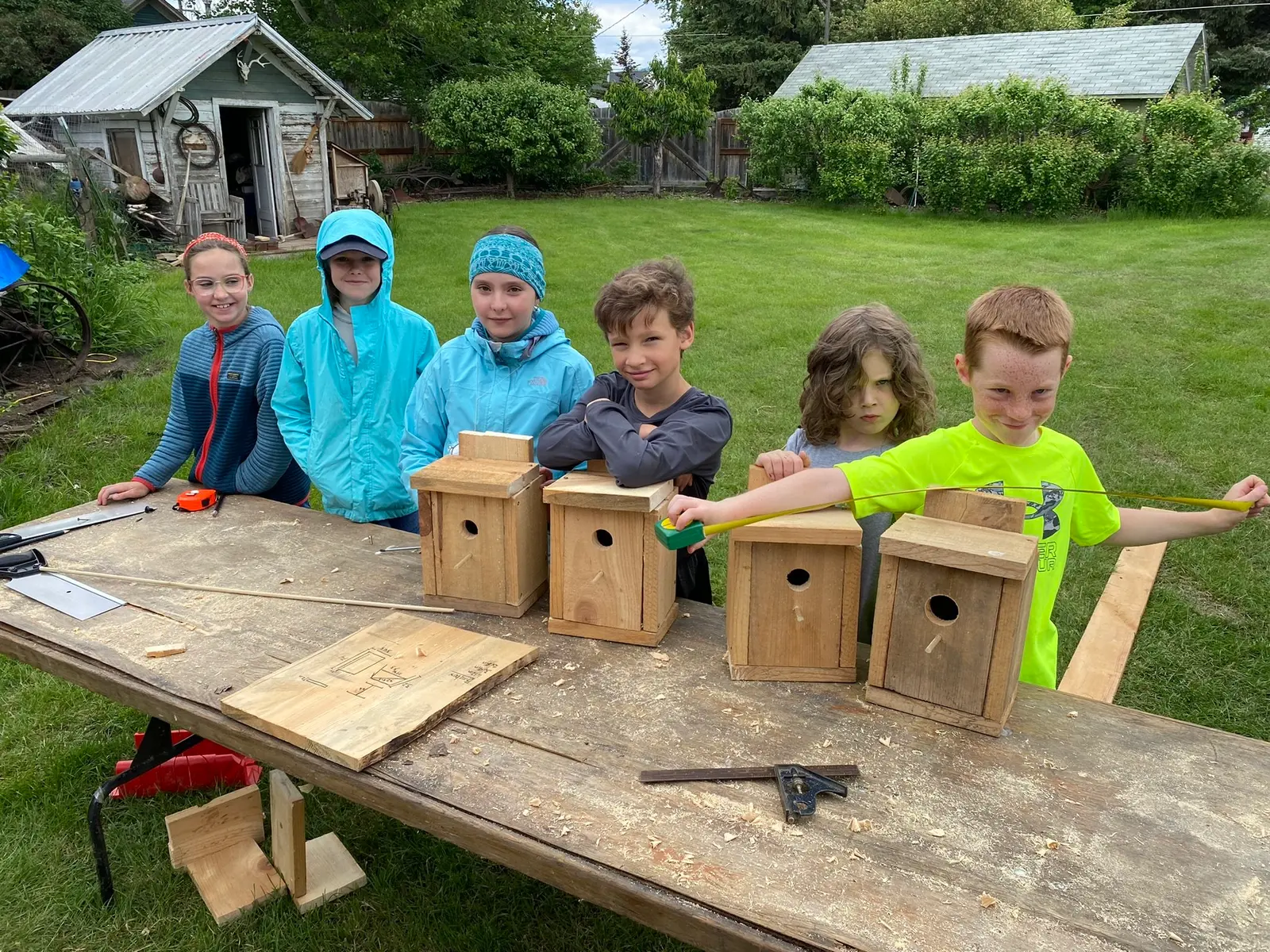 A group of children stand around a wooden table in a yard displaying their handcrafted birdhouses.