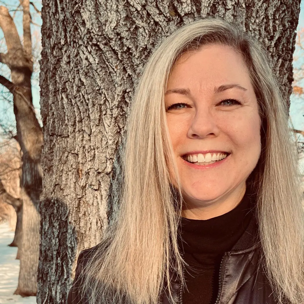 A smiling woman with blonde hair stands outside in front of a tree.