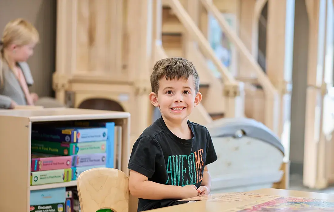 A young boy smiles while sitting at a table with puzzle boxes in the background.