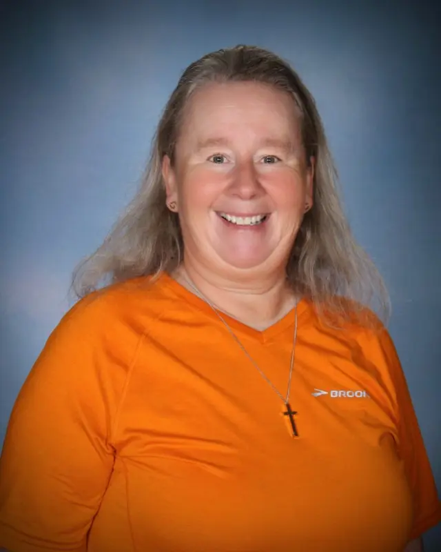A smiling woman wearing an orange athletic shirt and a cross necklace poses against a blue background.