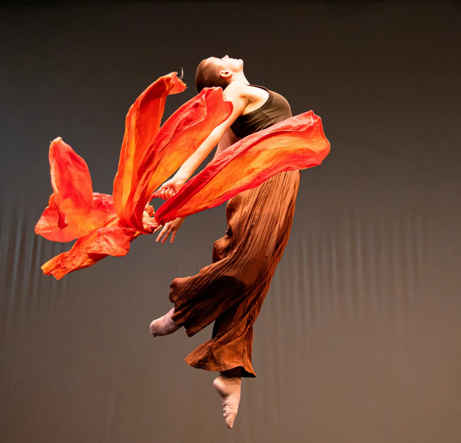 A dancer leaps through the air with flowing orange fabric against a dark background.