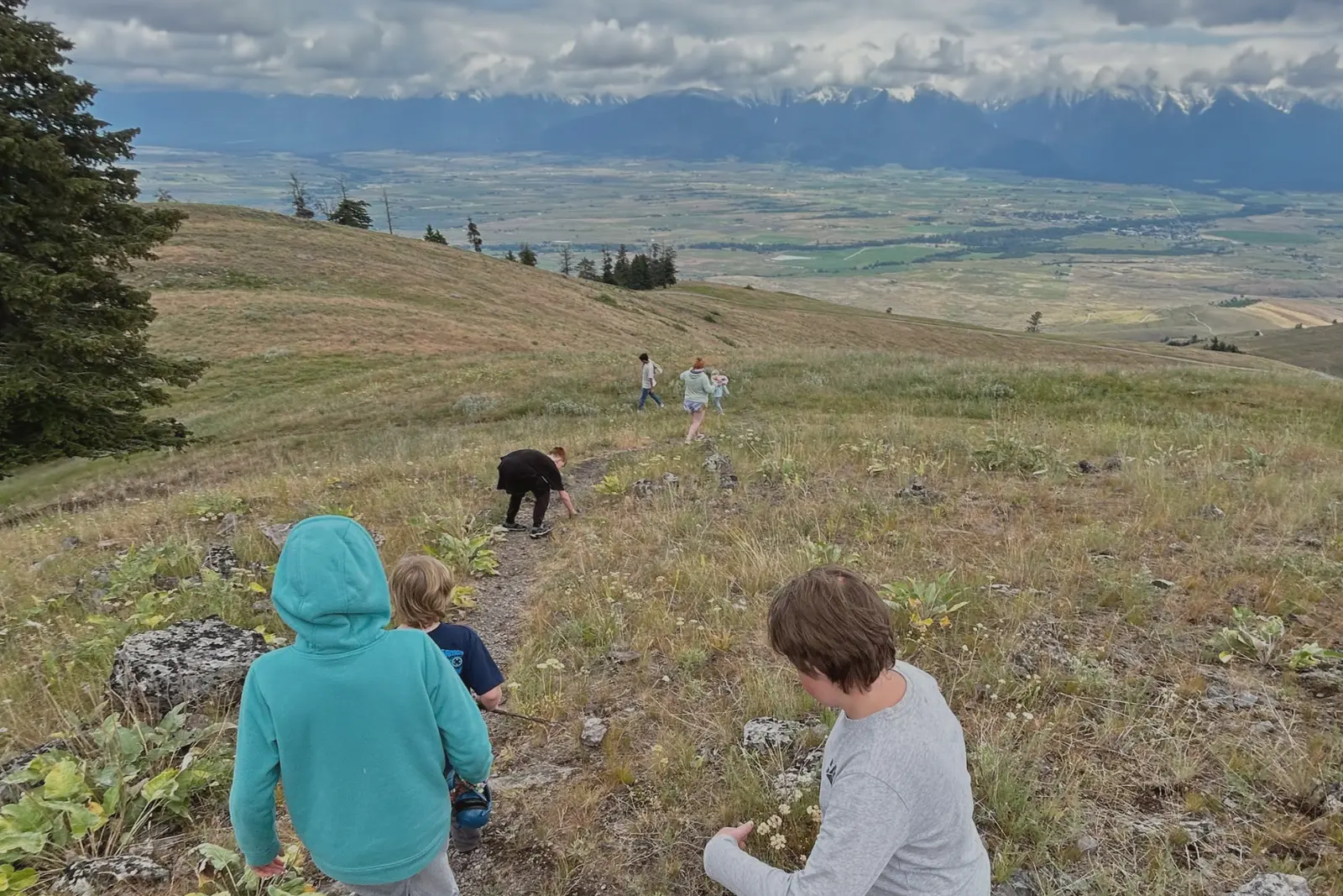 A group of young people hikes down a grassy hill overlooking a vast valley and distant mountains.