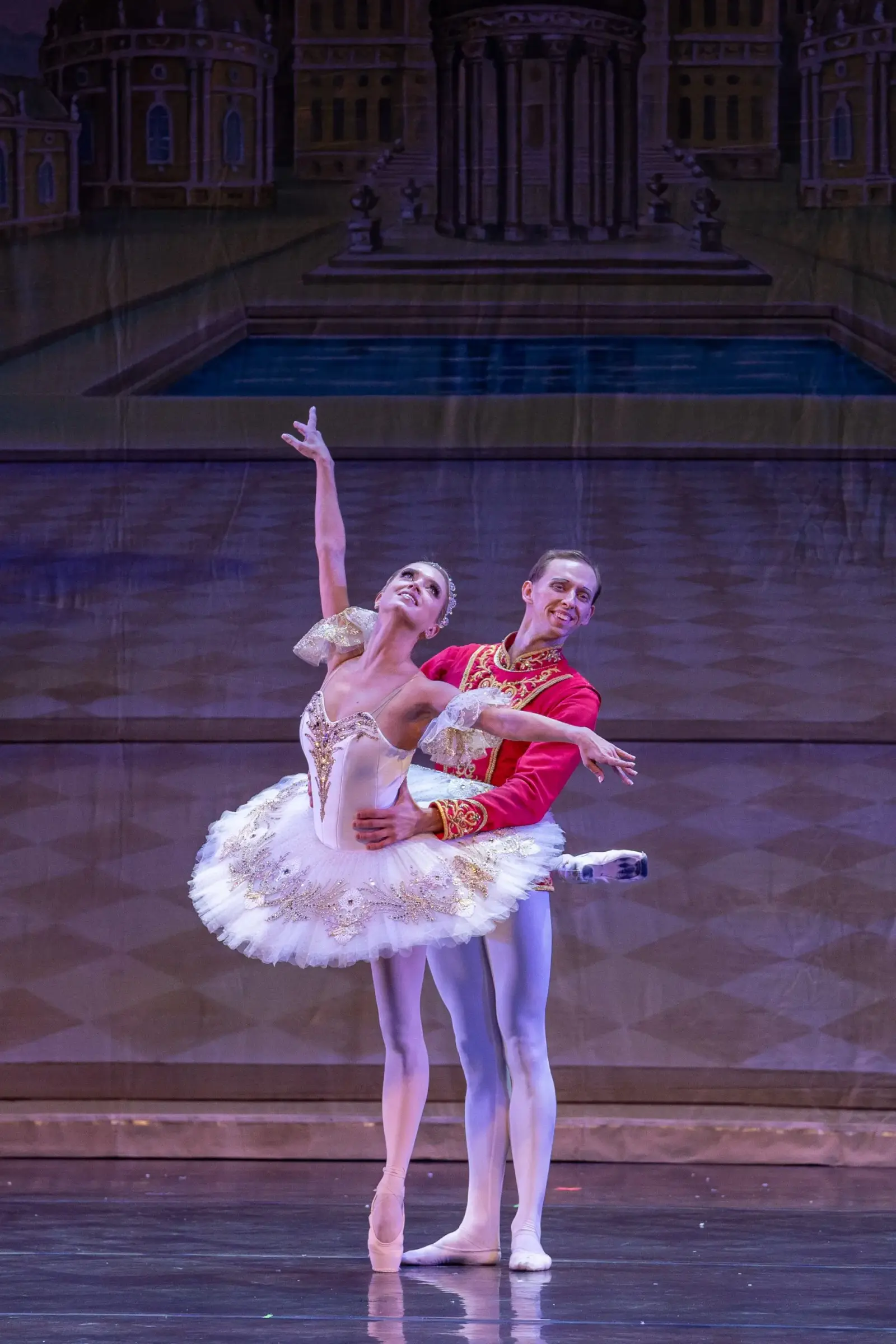 A male and female ballet dancer perform together on a stage in front of a painted backdrop.