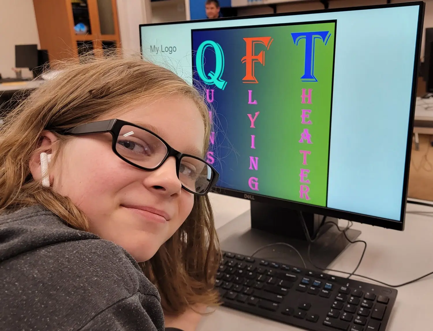A young person with glasses smiles while sitting in front of a computer screen displaying graphic text.