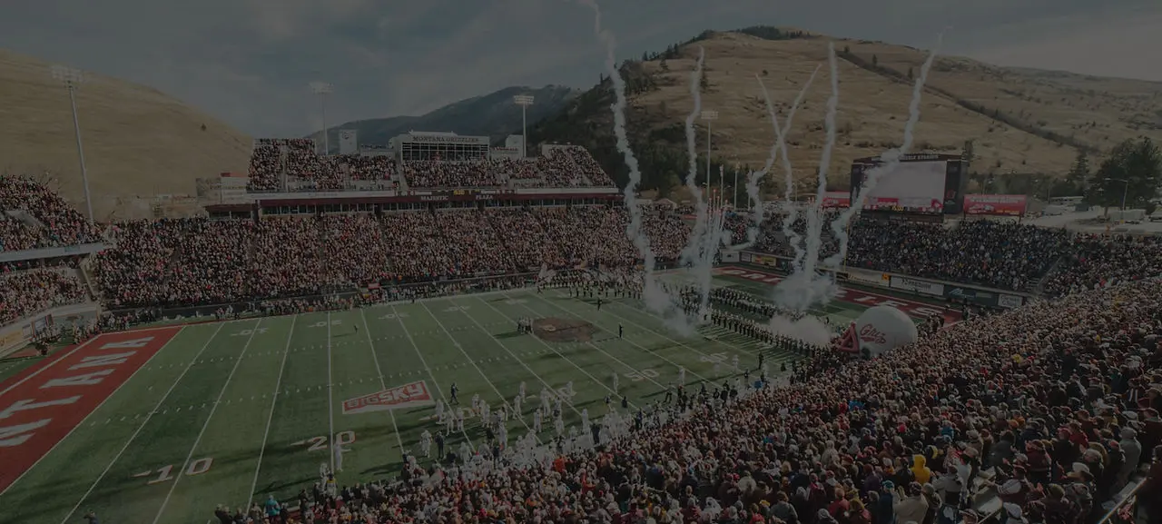 A large crowd fills a stadium for a football game as team players enter the field through smoke.