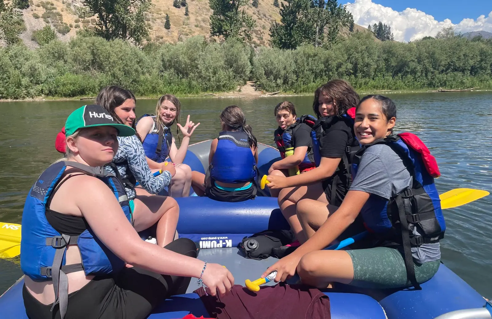 A group of children in life jackets sit in a raft on a river.