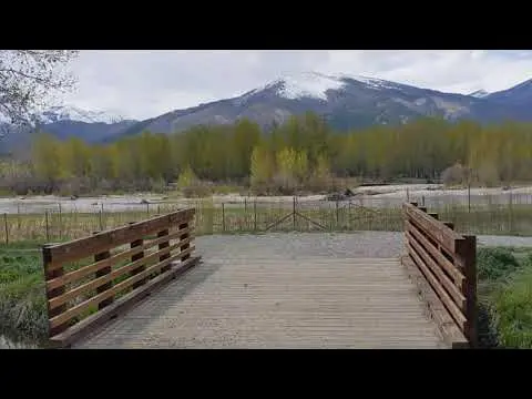 A wooden footbridge leads toward a scenic landscape with mountains and a river.