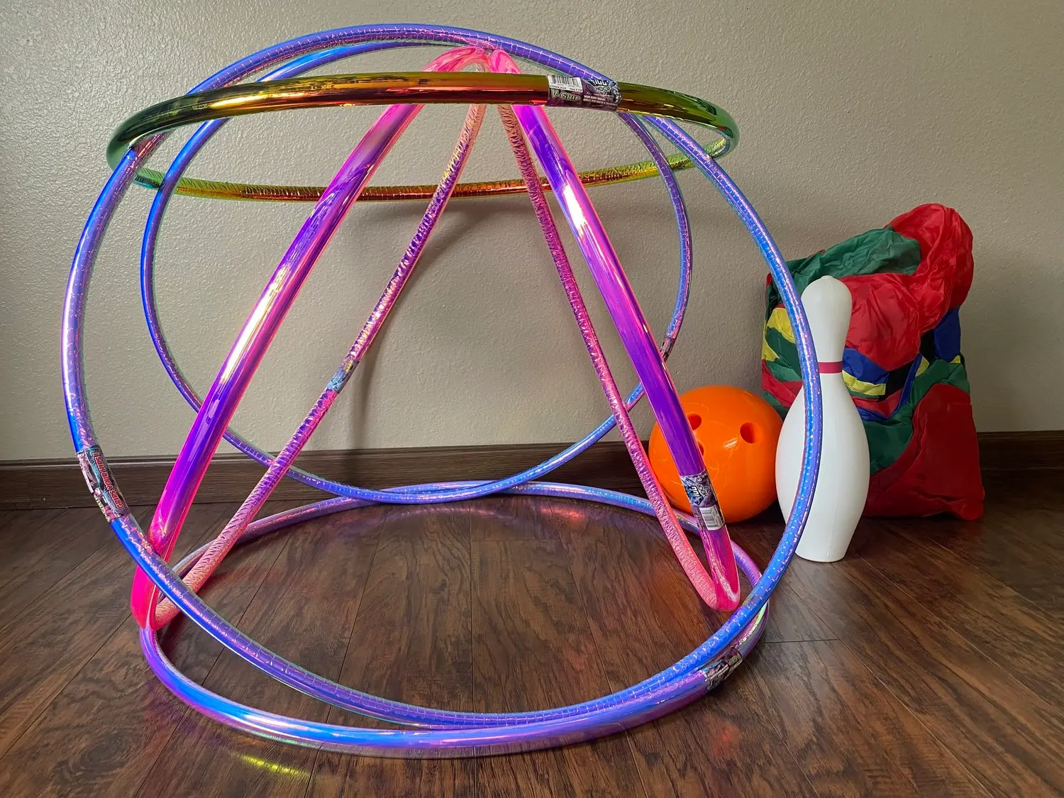 A collection of colorful hula hoops, a bowling pin, and an orange ball rest on a wood floor.