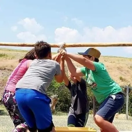 A group of children stand outside and hold their hands up to touch a horizontal wooden pole.